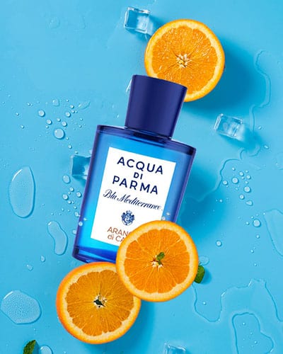 A bottle of Acqua Di Parma Blu Mediterraneo Arancia di Capri placed next to 3 ripe slices of orange fruit and some melting ice cubes, on a light blue background.