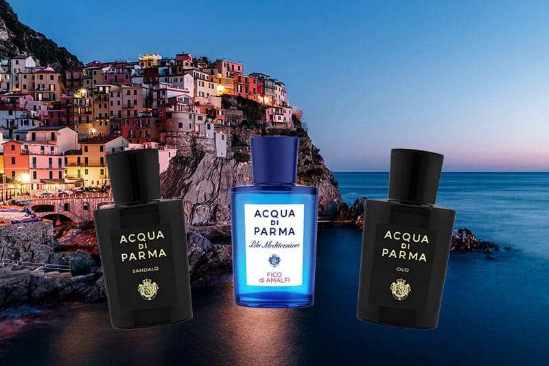 3 fragrance bottles in a row of Acqua Di Parma’s Sandalo, Fico Di Amalfi, and Oud with the Amalfi seaside town, cliffs, and coast in the background at dusk.