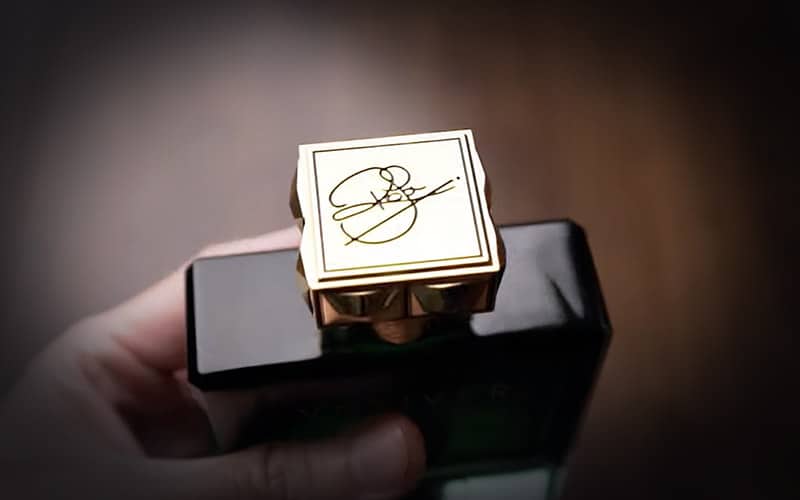 A close-up of Roja Dove’s signature engraved on the top of the cap of a bottle of Roja Parfums Vetiver Parfum Cologne, being held in one hand.