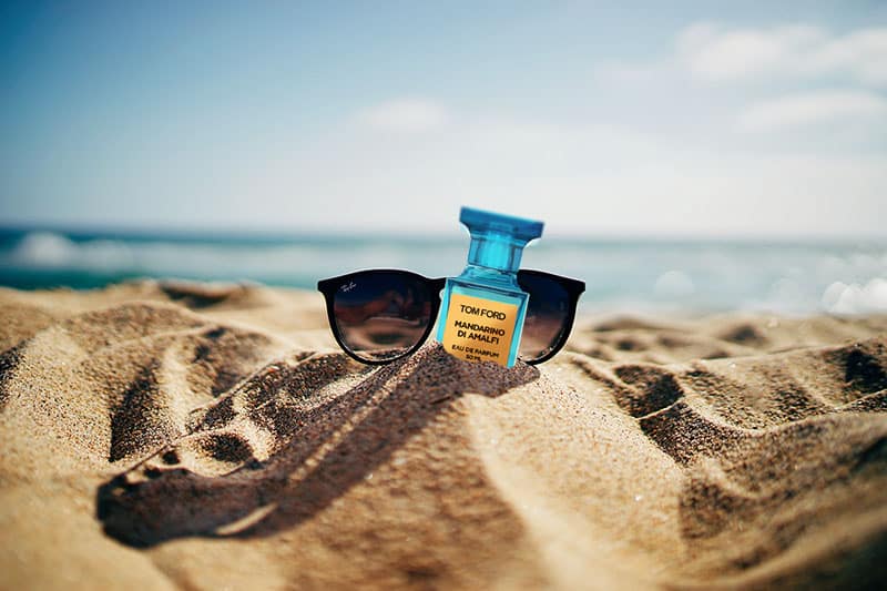 A bottle of Tom Ford Mandarino Di Amalfi in the sand next to a pair of sunglasses at the beach.