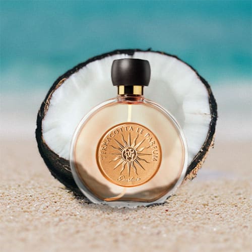 A bottle of Guerlain Terracotta Le Parfum partially buried in the sand on the beach, sitting directly in front of a fresh half-cut coconut shell with a sunny sea horizon in the background.