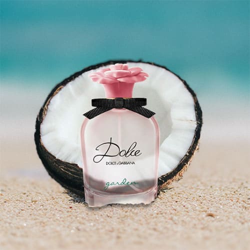 A bottle of Dolce and Gabbana Dolce Garden partially buried in the sand on the beach, sitting directly in front of a fresh half-cut coconut shell with a sunny sea horizon in the background.