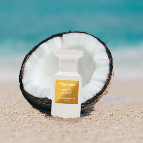 A bottle of Tom Ford Soleil Blanc partially buried in the sand on the beach, sitting directly in front of a fresh half-cut coconut shell with a sunny sea horizon in the background.