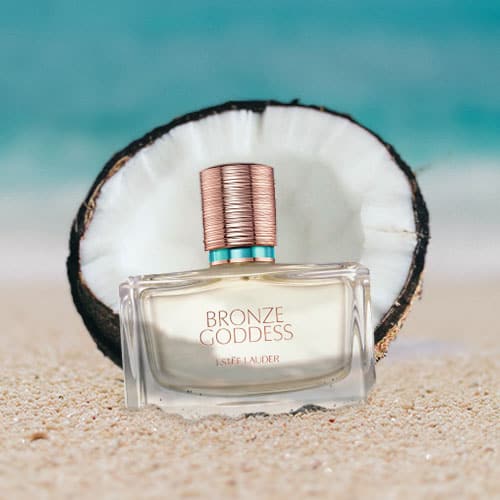 A bottle of Estee Lauder Bronze Goddess Eau Fraiche skinscent partially buried in the sand on the beach, sitting directly in front of a fresh half-cut coconut shell with a sunny sea horizon in the background.