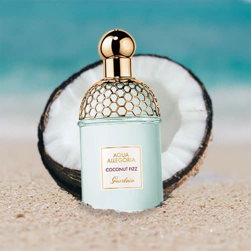 A bottle of Guerlain Coconut Fizz partially buried in the sand on the beach, sitting directly in front of a fresh half-cut coconut shell with a sunny sea horizon in the background.