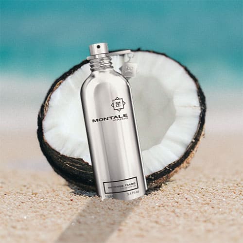A bottle of Montale Intense Tiare partially buried in the sand on the beach, sitting directly in front of a fresh half-cut coconut shell with a sunny sea horizon in the background.