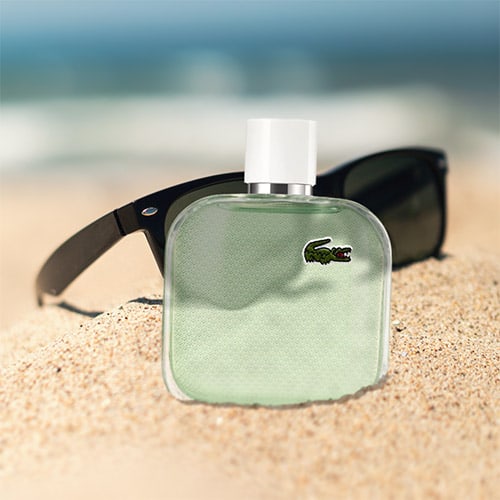 A bottle of Lacoste L1212 Blanc Eau Fraiche partially buried in sand on the beach, in front of a pair of sunglasses with the sea horizon in the background.