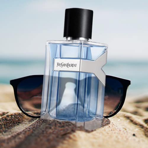 A bottle of YSL Y EDT partially buried in sand on the beach, in front of a pair of sunglasses with the sea horizon in the background.