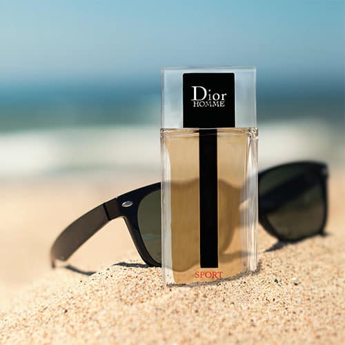 A bottle of Dior Homme Sport partially buried in sand on the beach, in front of a pair of sunglasses with the sea horizon in the background.