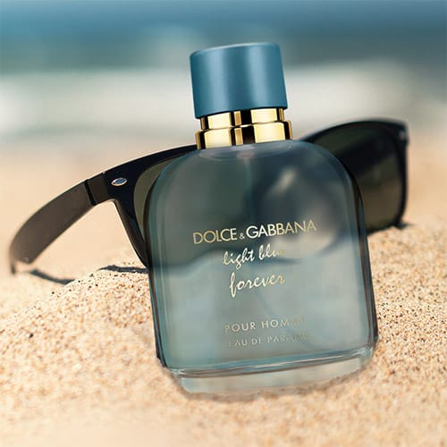 A bottle of Dolce & Gabbana Light Blue Forever Pour Homme partially buried in sand on the beach, in front of a pair of sunglasses with the sea horizon in the background.