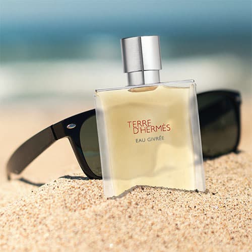 A bottle of Hermes Terre D’Hermes Eau Givree partially buried in sand on the beach, in front of a pair of sunglasses with the sea horizon in the background.