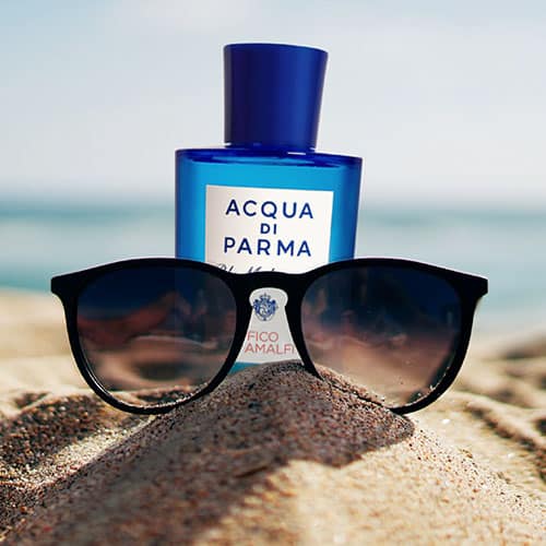 A bottle of Acqua Di Parma Fico Di Amalfi partially buried in sand on the beach, behind a pair of sunglasses with the sea horizon in the background.