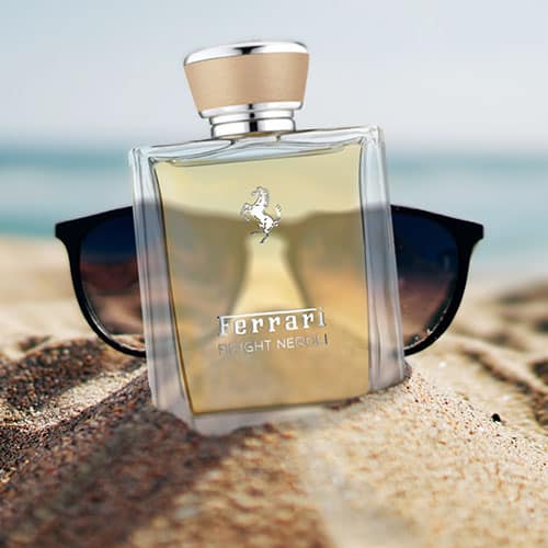A bottle of Ferrari Bright Neroli partially buried in sand on the beach, in front of a pair of sunglasses with the sea horizon in the background.