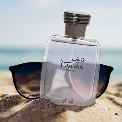 A bottle of Rasasi Hawas for Him partially buried in sand on the beach, in front of a pair of sunglasses with the sea horizon in the background.