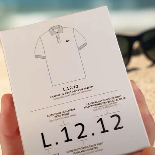 A close-up showing the information printed on the back of the packaging box of a bottle of Lacoste L.12.12 Blanc Eau Fraiche. The box is being held in one hand, with a pair of sunglasses in the sand and the sea horizon in the background.