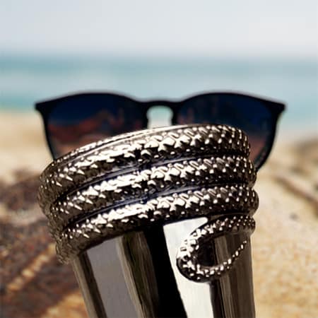 A close-up of the engraved python snake wrapped around the cap of a bottle of Rasasi Hawas for Him, with a pair of sunglasses in the sand and the sea horizon in the background.