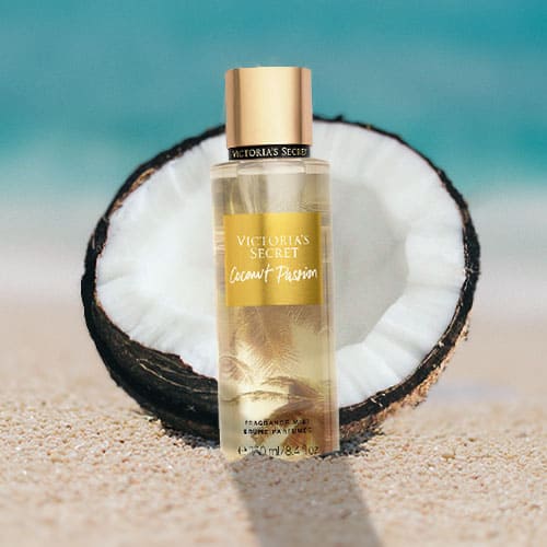 A bottle of Victoria’s Secret Coconut Passion partially buried in the sand on the beach, sitting directly in front of a fresh half-cut coconut shell with a sunny sea horizon in the background.