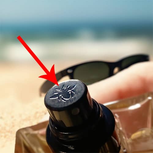 A red arrow pointing to a close-up of the Dior Homme bee logo engraved on top of the atomizer of a bottle of Dior Homme Sport, which is being held in one hand, with a pair of sunglasses and sea horizon in the background.