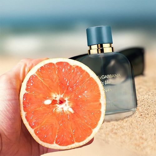 A freshly half-cut grapefruit being held in one hand. Behind the grapefruit is a bottle of Dolce & Gabbana Light Blue Forever Pour Homme and a pair of sunglasses, which are partially buried in sand on the beach, with the sea horizon in the background.