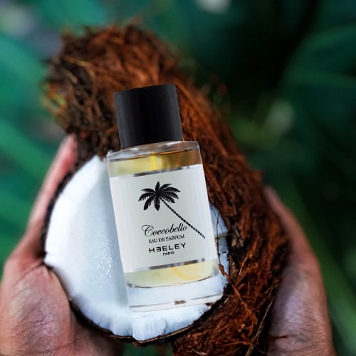 A bottle of Heeley Coccobello laid inside a fresh half-cut coconut shell. The half shell and removed husk are being held in the cupped palms of two hands with palm tree leaves in the background.