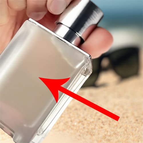 A red arrow pointing to a close-up of the frosted glass on the back of a bottle of Hermes Terre D’Hermes Eau Givree. The bottle is being held by its cap in one hand, in front of a pair of sunglasses in the sand, with the sea horizon in the background.