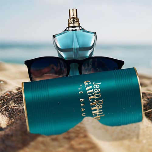 A bottle of Jean Paul Gaultier Le Beau and its cylindrical tin can, both partially buried in sand on the beach, next to a pair of sunglasses with the sea horizon in the background.