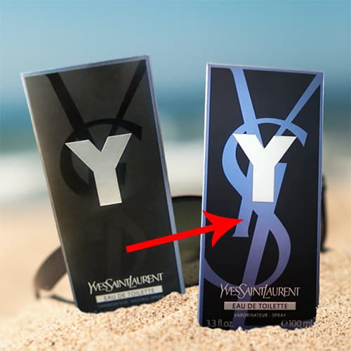 A packaging box of the previous formulation of YSL Y EDT next to the packaging box of the new formulation. Both boxes are partially buried in sand on the beach in front of a pair of sunglasses with the sea horizon in the background. And a red arrow points out the new blue colored YSL logo.