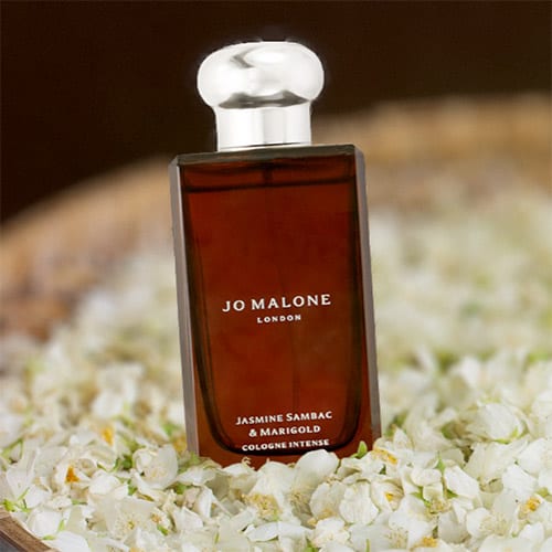A bottle of Jo Malone Jasmine Sambac & Marigold resting on a bed of jasmine flowers in a round woven basket.