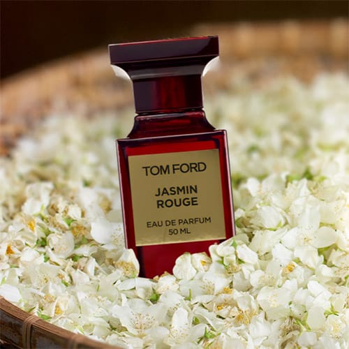 A bottle of Tom Ford Jasmin Rouge resting on a bed of jasmine flowers in a round woven basket.