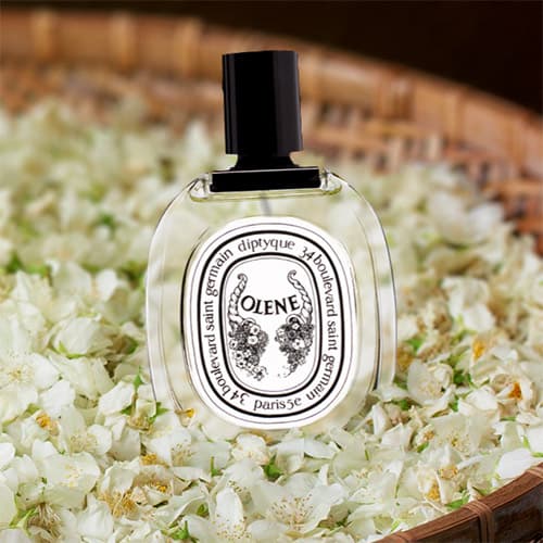 A bottle of Diptyque Olene resting on a bed of jasmine flowers in a round woven basket.