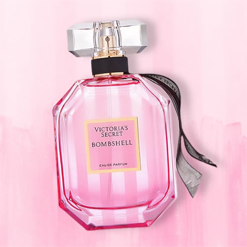 A bottle of Victoria’s Secret Bombshell EDP lying flat on top of a striped light pink colored table.