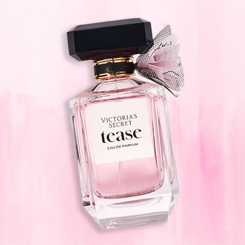 A bottle of Victoria’s Secret Tease EDP lying flat on top of a striped light pink colored table.