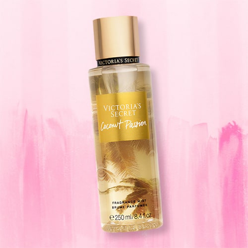 A bottle of Victoria’s Secret Coconut Passion Body Mist lying flat on top of a striped light pink colored table.
