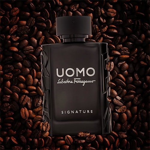 A bottle of Salvatore Ferragamo Uomo Signature placed faceup on top of a pile of dark roasted coffee beans.