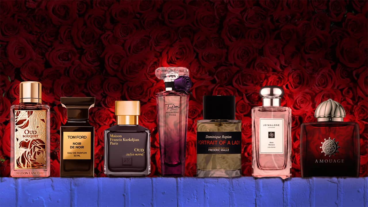 A selection of 7 bottles of the best rose perfumes from various brands. The bottles are lined up sitting on top of a pink brick wall. The bottles are individually illumined in the dim lighting with a dense wall of dark red roses in the background.