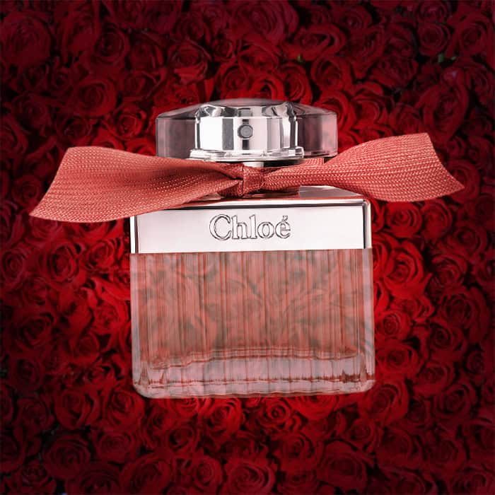 A close-up of an illuminated bottle of Chloe’s Roses De Chloe with a dense wall of dark red roses in the background.