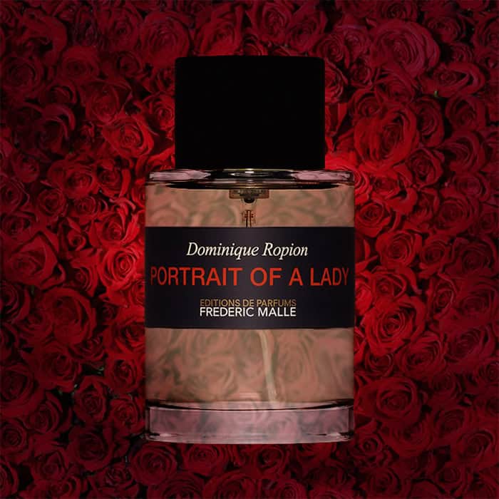 A close-up of an illuminated bottle of Frederic Malle’s Editions De Parfums Portrait of a Lady with a dense wall of dark red roses in the background.