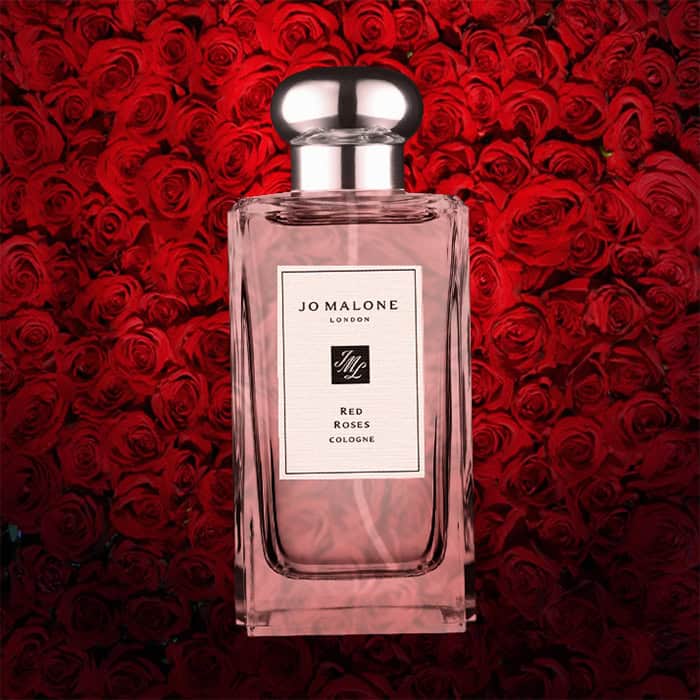 A close-up of an illuminated bottle of Jo Malone Red Roses Cologne with a dense wall of dark red roses in the background.