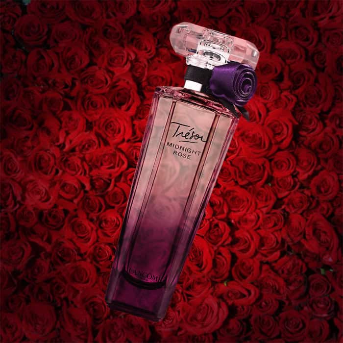 A close-up of an illuminated bottle of Lancome Tresor Midnight Rose with a dense wall of dark red roses in the background.