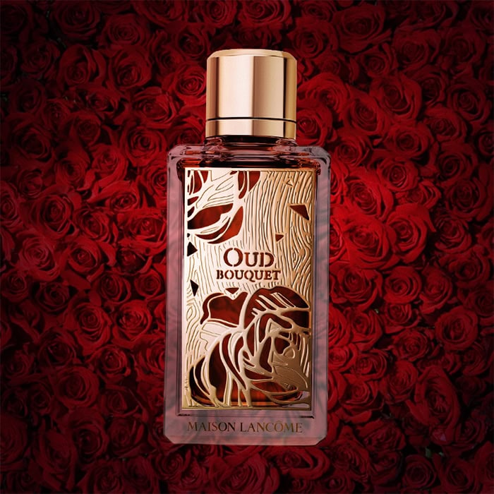 A close-up of an illuminated bottle of Maison Lancome Oud Bouquet with a dense wall of dark red roses in the background.