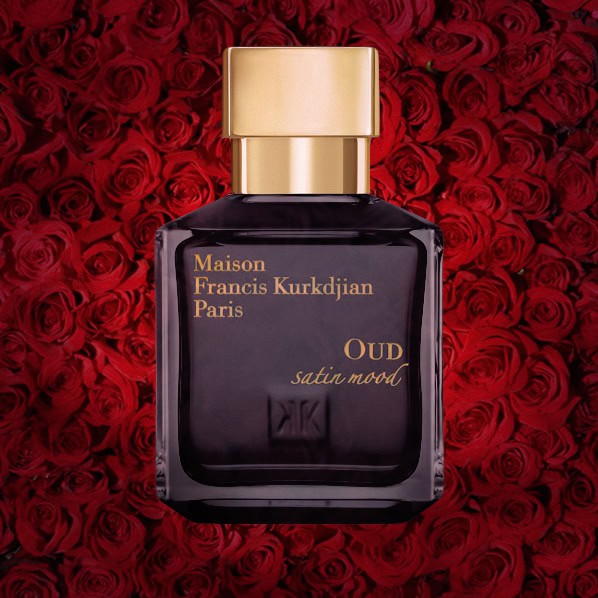 A close-up of an illuminated bottle of MFK Maison Francis Kurkdjian Oud Satin Mood with a dense wall of dark red roses in the background.