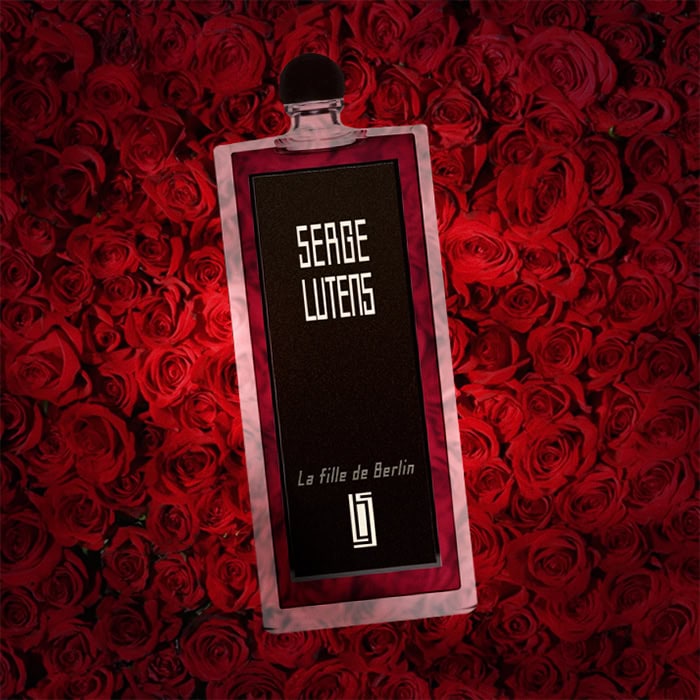 A close-up of an illuminated bottle of Serge Lutens La Fille de Berlin with a dense wall of dark red roses in the background.