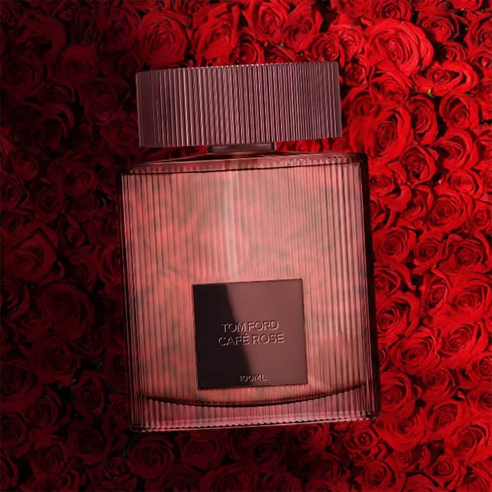 A close-up of an illuminated bottle of Tom Ford Cafe Rose with a dense wall of dark red roses in the background.