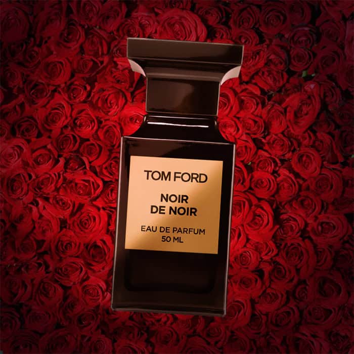 A close-up of an illuminated bottle of Tom Ford Noir De Noir with a dense wall of dark red roses in the background.