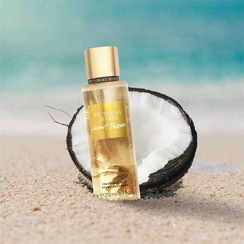 A bottle of Victoria’s Secret Coconut Passion partially buried in the sand on the beach, sitting directly in front of a fresh half-cut coconut shell with a sunny sea horizon in the background.