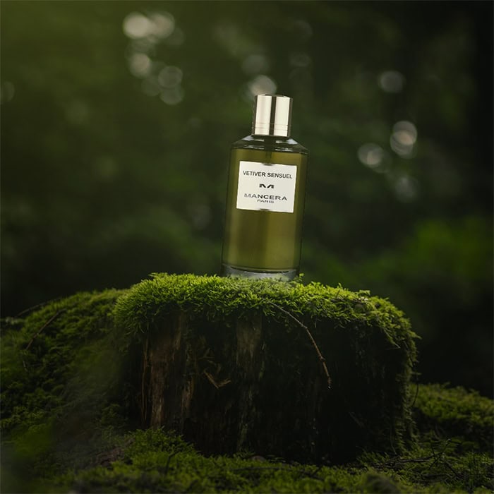 A close-up of a bottle of Mancera Vetiver Sensuel sitting on top of an old green moss-covered tree stump in the forest.