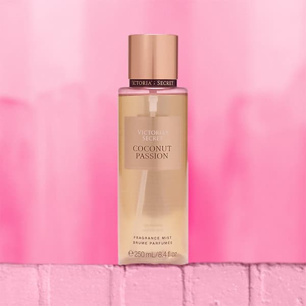 A close-up of the new, updated Victoria’s Secret Coconut Passion Body Mist bottle. The bottle is sitting on top of a light pink colored brick wall.