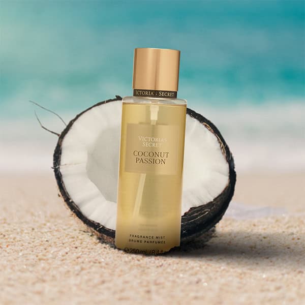 A close-up of the new, updated Victoria’s Secret Coconut Passion body mist bottle. The bottle is partially buried in the sand on the beach, and is sitting directly in front of a fresh half-cut coconut shell with a sunny sea horizon in the background.