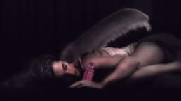 A woman lying asleep on her side in a dimly-lit room. She is wearing a white angel wings costume, and sitting next to her left hand is the new, updated Victoria’s Secret Very Sexy Eau De Parfum bottle. The bottle emits a soft red glow, which partially softy lights her face and arms.