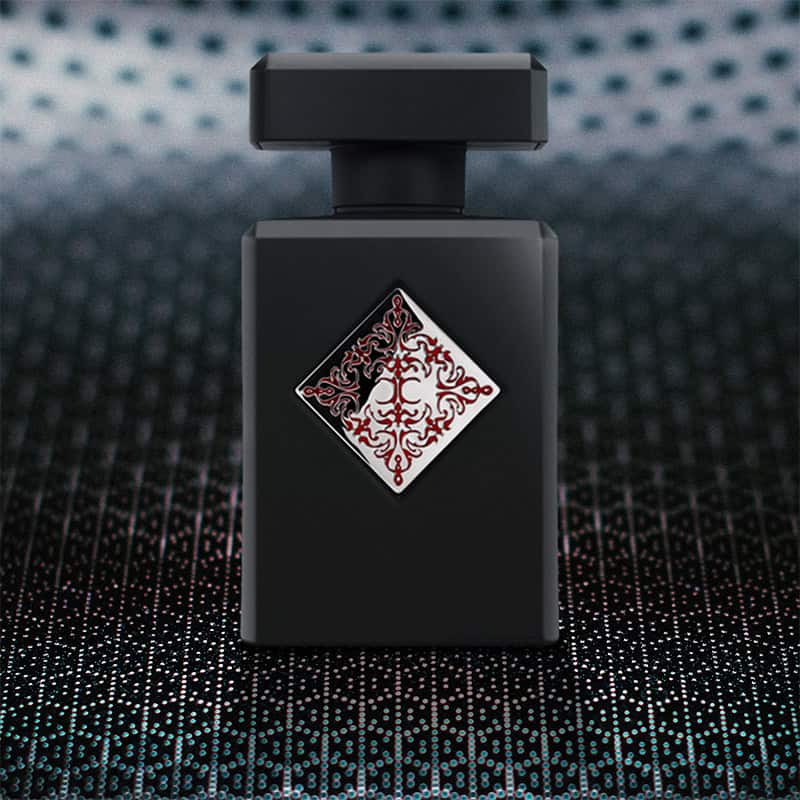 A close-up of the front face of a bottle of Initio Parfums Absolute Aphrodisiac. The bottle is sitting on a textured grey-colored hexagonal-patterned surface and there is a blurred hexagonal-patterned background.